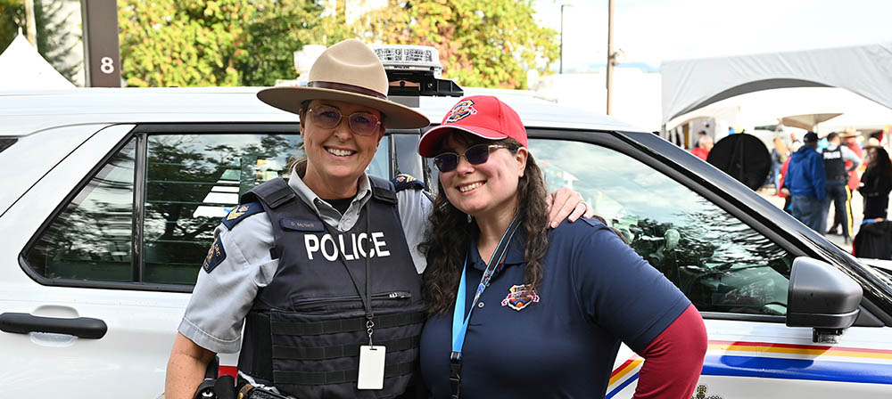 Une policière souriante vêtue d’un uniforme de la GRC et d’un stetson se tient devant un VUS de la police à l’extérieur. Elle entoure de son bras une employée souriante de la Ville de Burnaby qui porte un t-shirt bleu, une casquette rouge et des lunettes de soleil.