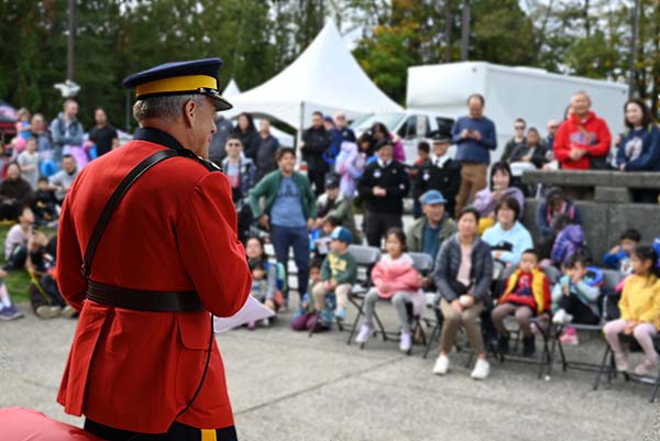 A police office in Red Serge speaks into a microphone in front of a crowd of people on a cloudy day