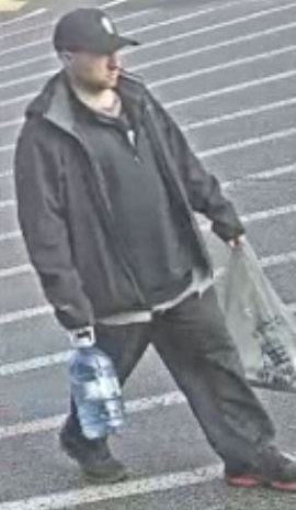 Caucasian man walking through a parking lot, wearing a black baseball cap, dark sweater, dark jacket, jeans and black shoes.