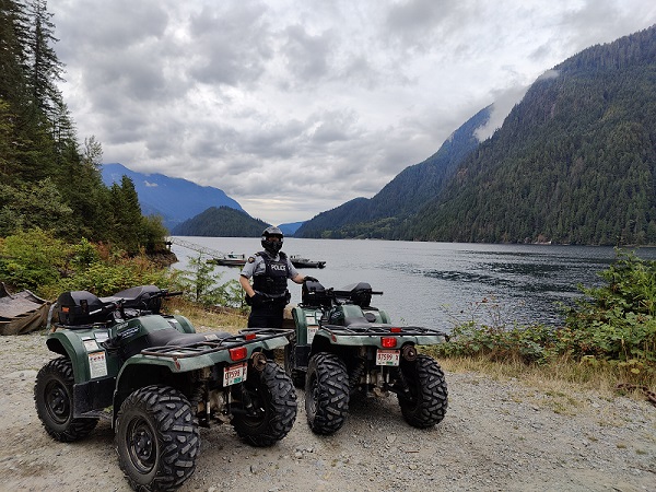 Photo d’un membre de la GRC portant un casque intégral, debout devant deux VTT avec une vue sur une zone forestière, un lac et des montagnes en arrière-plan.