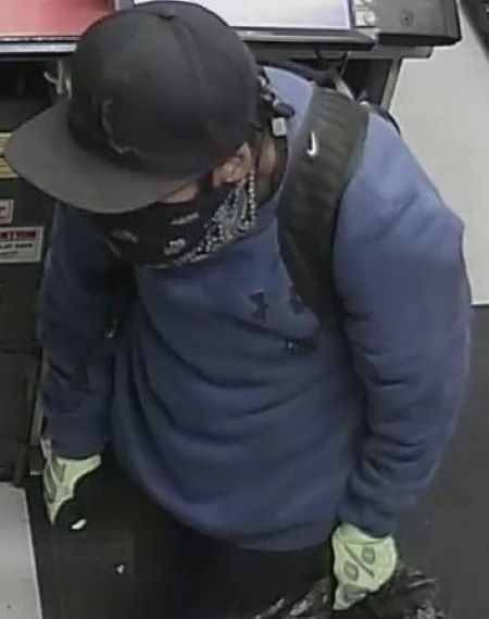 Suspect blue sweater: A suspect in a blue sweater, greenish gloves, black hair, and dark bandana covering his face. A black backpack strap with a white Nike symbol is visible on his shoulder.
