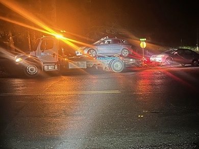 A tow truck has emergency lights flashing and is towing two grey vehicles